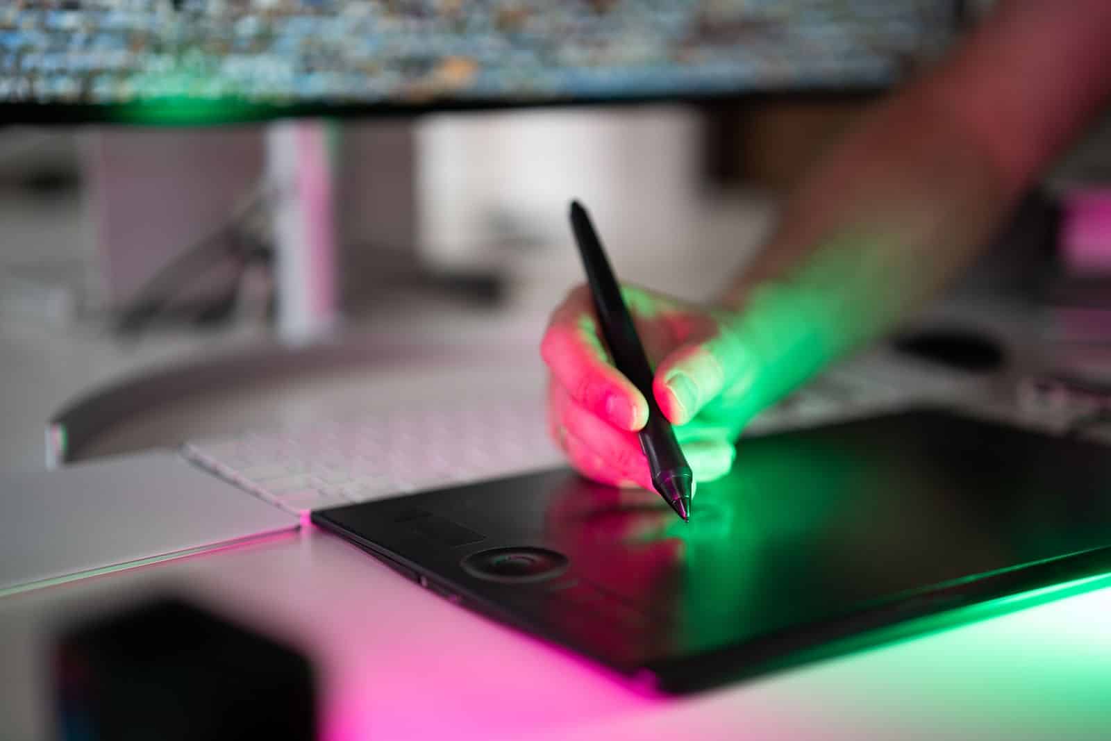 Hand using a stylus on a graphics tablet illuminated by colorful pink and green light, representing digital art creation